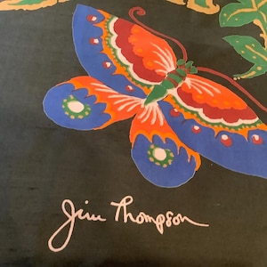 Puede incluir: Una tela negra con una mariposa grande y colorida de alas azules, naranjas, rojas y verdes. La mariposa est&aacute; rodeada de hojas verdes y elementos florales naranjas. El nombre Jim Thompson aparece impreso en letra blanca en la parte inferior.