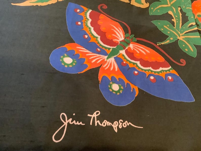 Puede incluir: Una tela negra con una mariposa grande y colorida de alas azules, naranjas, rojas y verdes. La mariposa est&aacute; rodeada de hojas verdes y elementos florales naranjas. El nombre Jim Thompson aparece impreso en letra blanca en la parte inferior.