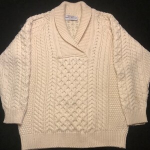 Peut inclure: Pull en maille crème avec un col châle. Le pull présente divers motifs texturés, notamment des tresses et des losanges. L'étiquette indique "Royal County of Dublin, Margaret O'Neill Ireland".