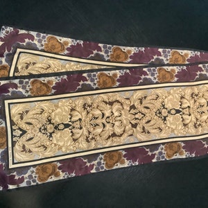 May include: A long, rectangular silk scarf with a floral pattern. The scarf features a repeating design of brown, purple, and blue flowers on a light blue background. The centre of the scarf has a gold and brown damask pattern.