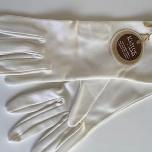 Peut inclure: Une paire de gants blancs arrivant au poignet avec une finition lisse. Une étiquette ronde en papier marron est attachée au poignet avec un cordon doré, portant le texte Kidtex by Kayser, indiquant que ces gants en nylon imitent le cuir.
