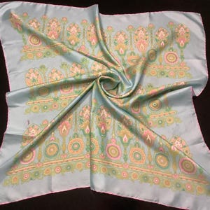 Peut inclure: Un foulard carré en soie bleu clair avec un motif floral répété en rose, vert et jaune. Le foulard est plié et drapé, mettant en valeur le motif détaillé et le brillant du tissu. Les bords sont finis avec une bordure rose.