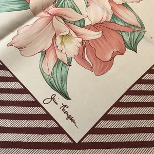May include: A white background with a brown and white striped border.  The centre of the background features a pink and white floral design with green leaves.  The signature "Gin Thompson" is written in black ink in the bottom right corner.