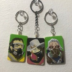 May include: Three painted wooden keychains featuring characters from a video game. The keychains are green, yellow, and brown with black and white details. The characters have masks and goggles.
