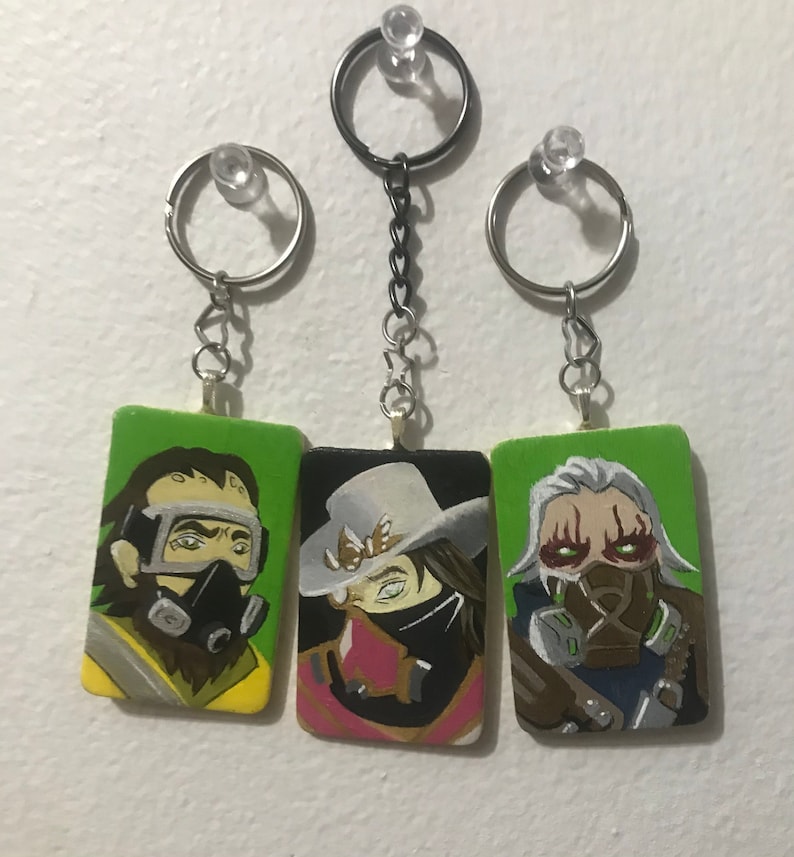 May include: Three painted wooden keychains featuring characters from a video game. The keychains are green, yellow, and brown with black and white details. The characters have masks and goggles.