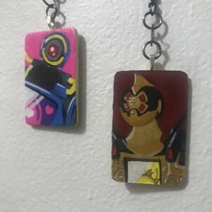 May include: Two painted wooden keychains with metal rings. The keychain on the left is pink, blue, and yellow with a cartoon robot. The keychain on the right is red, brown, and yellow with a cartoon creature with a red eye.