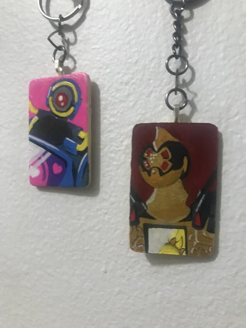 May include: Two painted wooden keychains with metal rings. The keychain on the left is pink, blue, and yellow with a cartoon robot. The keychain on the right is red, brown, and yellow with a cartoon creature with a red eye.