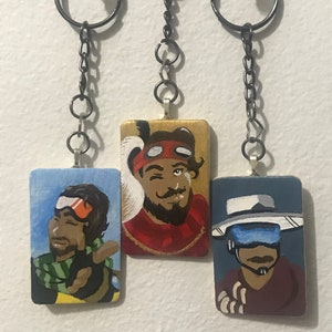 May include: Three painted wooden keychains featuring characters from the video game Apex Legends. The keychains are on a white background. The first keychain features a character with a green scarf and orange goggles. The second keychain features a character with a red bandana and a beard. The third keychain features a character with a white hat and a blue visor.