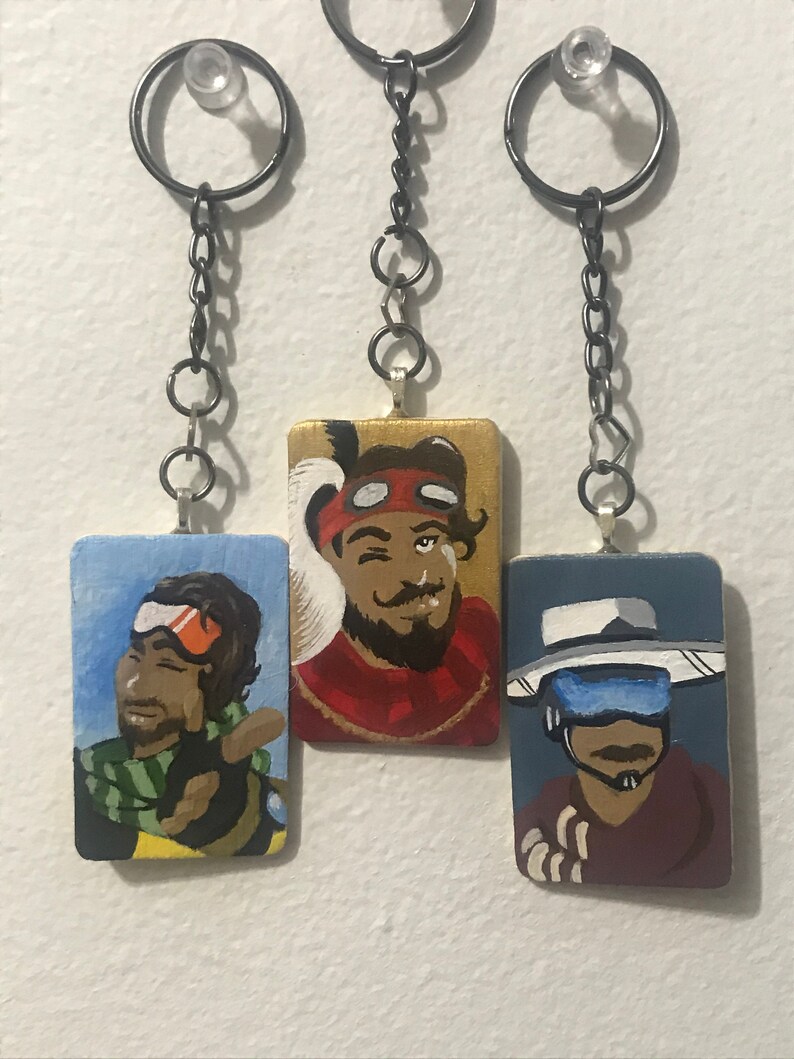 May include: Three painted wooden keychains featuring characters from the video game Apex Legends. The keychains are on a white background. The first keychain features a character with a green scarf and orange goggles. The second keychain features a character with a red bandana and a beard. The third keychain features a character with a white hat and a blue visor.