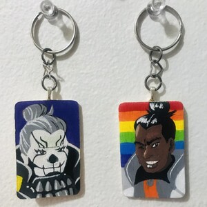 May include: Two keychains with painted wooden charms. One charm features a blue and white character with a skull-like face. The other charm features a rainbow background with a brown-skinned character with a black afro.