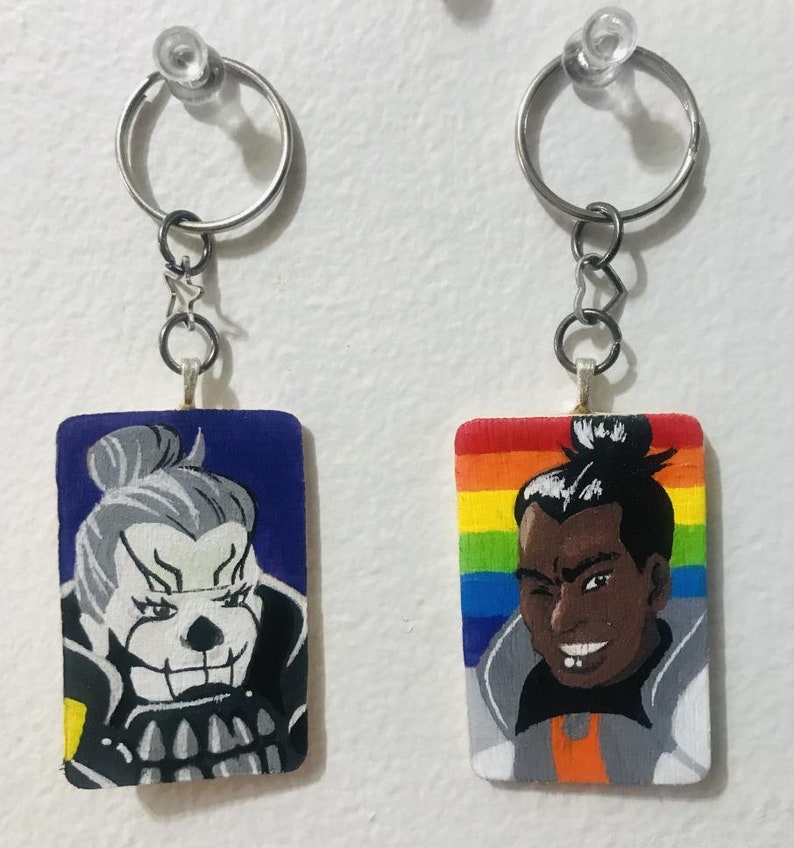 May include: Two keychains with painted wooden charms. One charm features a blue and white character with a skull-like face. The other charm features a rainbow background with a brown-skinned character with a black afro.