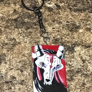 May include: A black and white painted wooden keychain with a red background. The keychain features a skull with horns and a detailed design.