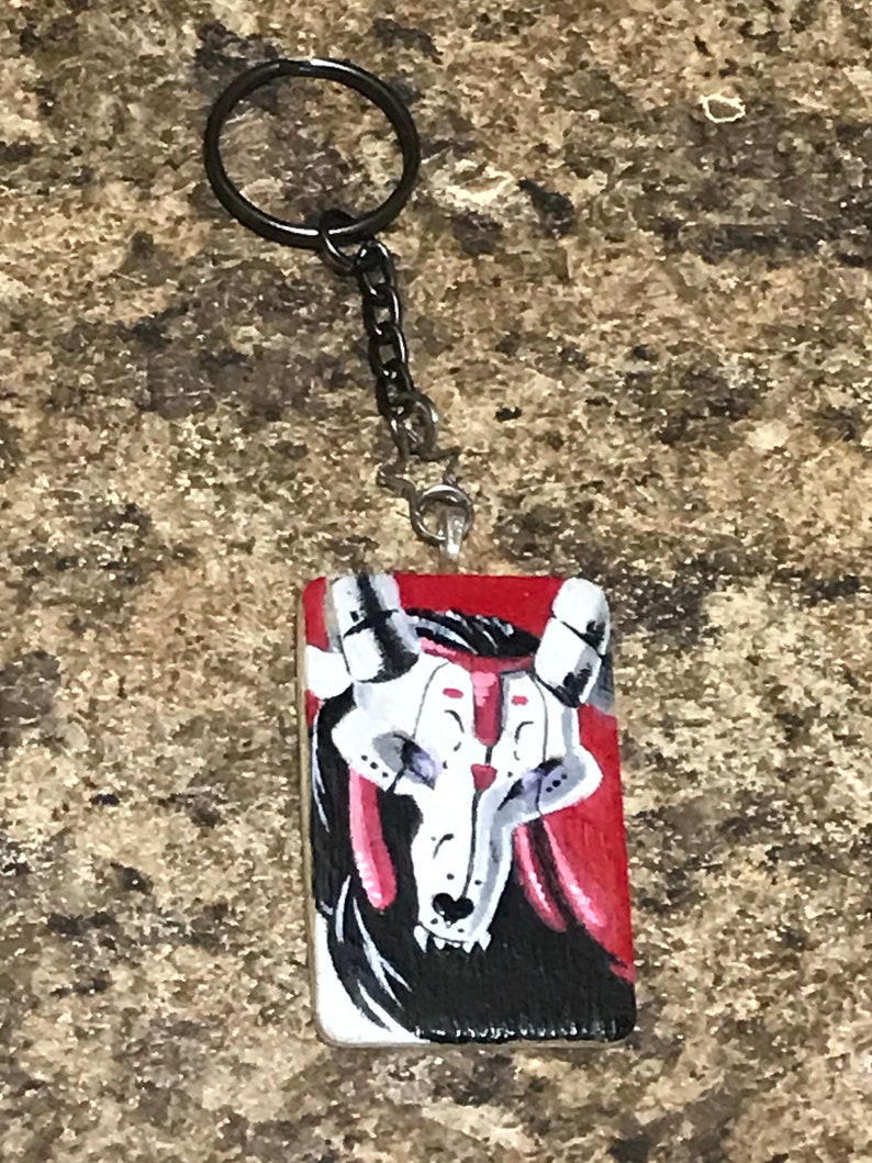 May include: A black and white painted wooden keychain with a red background. The keychain features a skull with horns and a detailed design.