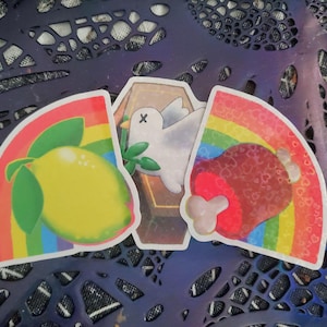 May include: Three die-cut stickers featuring a yellow lemon with green leaves, a white dove with a green branch inside a brown coffin, and a red meat bone, all set against colourful rainbow backgrounds with a subtle heart pattern on the meat sticker.