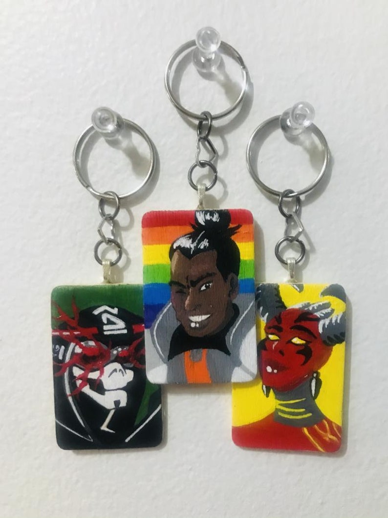May include: Three keychains with painted wooden charms. The charms feature a rainbow, a person with a black bun, and a red demon. The keychains are attached to silver rings.