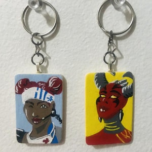 May include: Two painted wooden keyrings with metal rings. The keyring on the left features a portrait of a woman with red hair and a blue and white headband. The keyring on the right features a portrait of a red-skinned person with horns and yellow eyes.