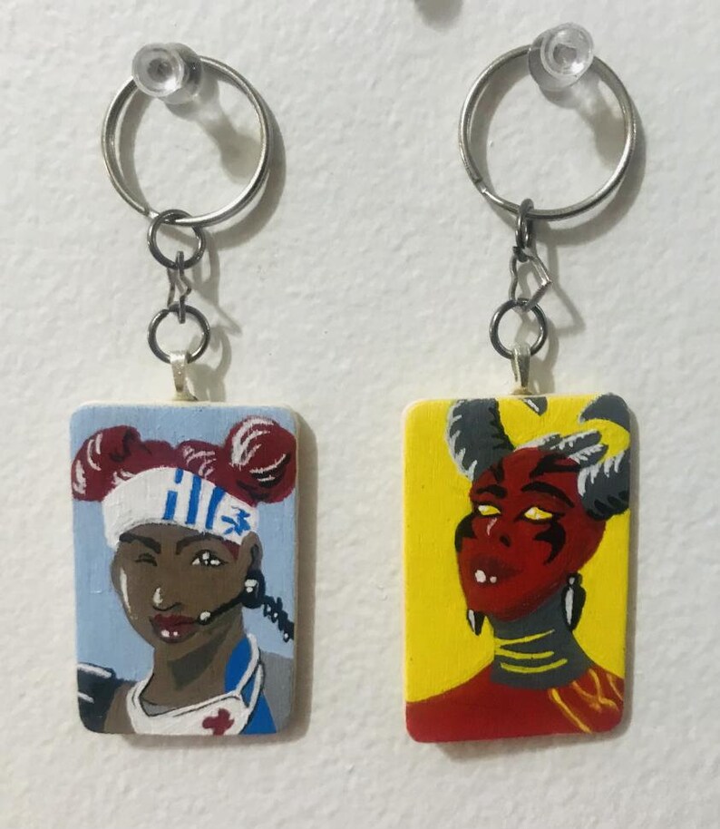 May include: Two painted wooden keyrings with metal rings. The keyring on the left features a portrait of a woman with red hair and a blue and white headband. The keyring on the right features a portrait of a red-skinned person with horns and yellow eyes.