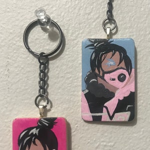 May include: Two painted wooden keychains featuring a character with black hair and a pink mask. The keychain on the left has a pink background and the keychain on the right has a blue background.