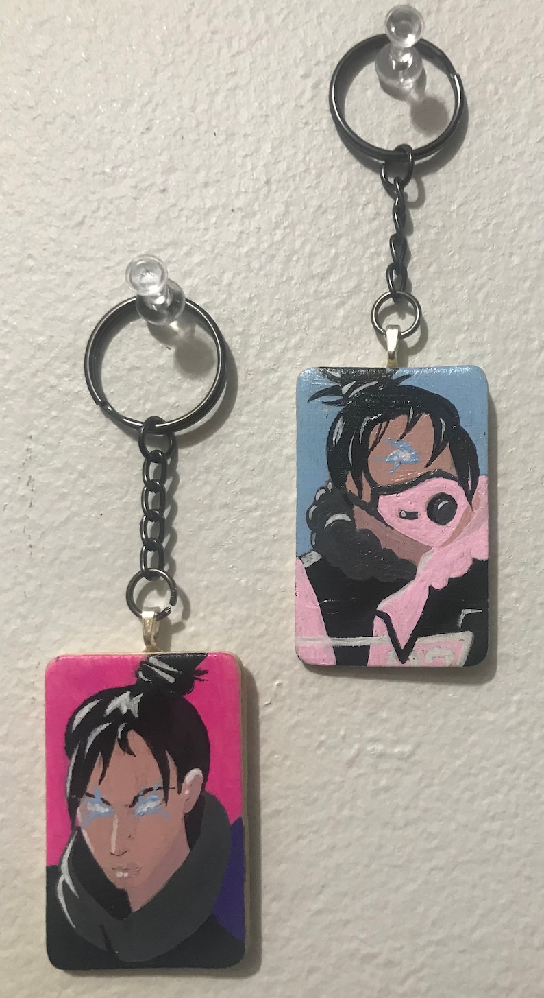 May include: Two painted wooden keychains featuring a character with black hair and a pink mask. The keychain on the left has a pink background and the keychain on the right has a blue background.