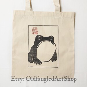 May include: Cream canvas tote bag with a black and white frog print in a rectangular border. The frog has a large open mouth. The tote bag has two handles and the text "Etsy: OldfangledArtShop" at the bottom.