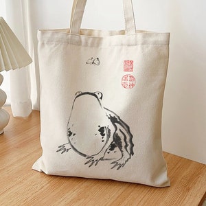 May include: A cream-colored tote bag featuring a black and white frog illustration. The frog is drawn in a minimalist style with a butterfly and red stamps. The bag has long handles for carrying.