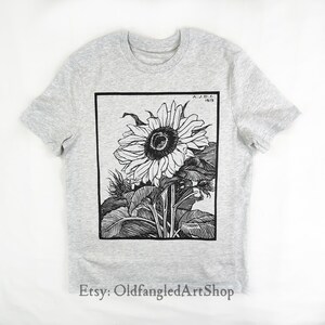 K&ouml;nnte beinhalten: Hellgraues T-Shirt mit einer Schwarz-Wei&szlig;-Illustration einer Sonnenblume. Das Kunstwerk ist in einem schwarzen Rechteck gerahmt und enth&auml;lt den Text "A.J.D.C. 1919". Der Etsy-Shop-Name "OldfangledArtShop" befindet sich unten.