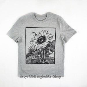 K&ouml;nnte beinhalten: Ein graues T-Shirt mit einer schwarz-wei&szlig;en Sonnenblumenillustration in einem schwarzen rechteckigen Rahmen. Der Etsy-Shop-Name "OldfangledArtShop" ist unten aufgedruckt. Das T-Shirt hat kurze &Auml;rmel und einen Rundhalsausschnitt.