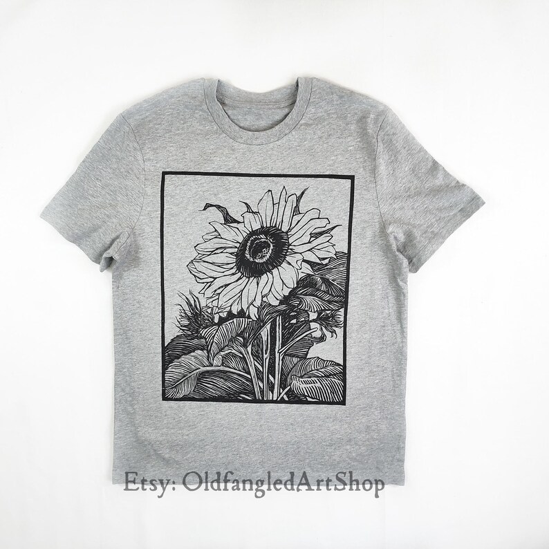 K&ouml;nnte beinhalten: Ein graues T-Shirt mit einer schwarz-wei&szlig;en Sonnenblumenillustration in einem schwarzen rechteckigen Rahmen. Der Etsy-Shop-Name "OldfangledArtShop" ist unten aufgedruckt. Das T-Shirt hat kurze &Auml;rmel und einen Rundhalsausschnitt.