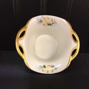 May include: A small, white porcelain dish with gold trim and floral designs. The dish has two handles and is perfect for serving small snacks or desserts.