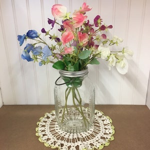 May include: A bouquet of pink, white and blue flowers in a clear glass jar with a silver lid. The jar is sitting on a white crocheted doily.