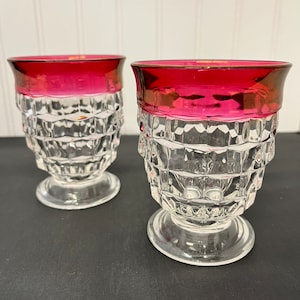 May include: Two clear glass tumblers with a red, ruby-colored rim. The glasses have a diamond-shaped pattern and a short, flared base.
