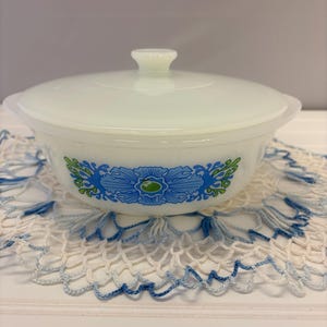 May include: A white milk glass casserole dish with a matching lid, featuring a decorative blue and green floral pattern on the side. The bowl has integrated side handles and a central knob on the lid, showcasing a symmetrical design.