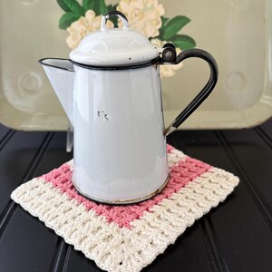 May include: A white enamel coffee pot with a black handle, a hinged lid, and a black rim, resting on a square crochet coaster featuring alternating bands of cream and pink yarn. The pot has a small spout and a metallic base rim.