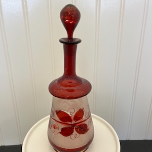 May include: A red glass decanter with a white floral design. The decanter has a round body and a tall, slender neck with a stopper.