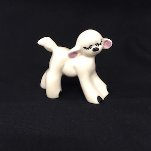 May include: White ceramic figurine of a lamb with closed eyes, pink ears, and black hooves. The lamb is standing on a black surface.