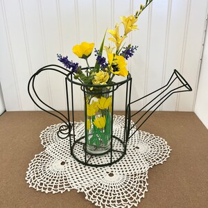 May include: A black metal watering can vase with a glass cylinder inside. The vase is filled with yellow and purple flowers. The vase is sitting on a white crocheted doily.
