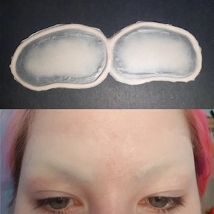 May include: A pair of white silicone eyebrow stencils, designed to help create symmetrical eyebrows. The stencils are shaped like a pair of eyebrows, with a curved shape and a defined arch.