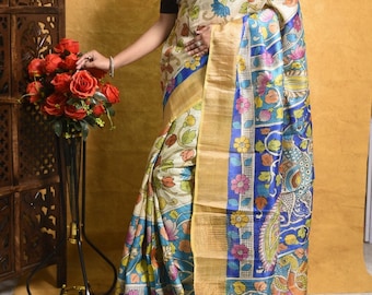 May include: Cream-colored sari with a floral and peacock design. The sari features a blue border with floral patterns and a gold-colored trim. The floral patterns include pink, blue, and yellow flowers. The sari is draped over a person.