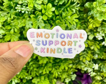 May include: A white sticker with a rainbow-colored cloud shape. The sticker says "Emotional Support Kindle" with a small flower graphic.