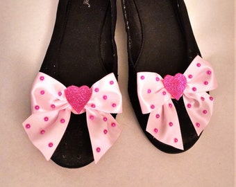 May include: A pair of black ballet flats with pink satin bows adorned with pink polka dots and a pink glitter heart in the center of each bow. The shoes are labeled "Wanted".