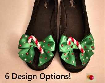 May include: A pair of black flats featuring green satin bows adorned with small red and white beads and a miniature candy cane decoration on each toe. The interior of the shoes displays the brand name Wanted. The text 6 Design Options! is visible.