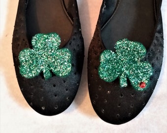 May include: A pair of black flats with green glitter shamrock embellishments. The shoes are decorated with small holes and the brand name "Wanted" is visible on the inside of each shoe.