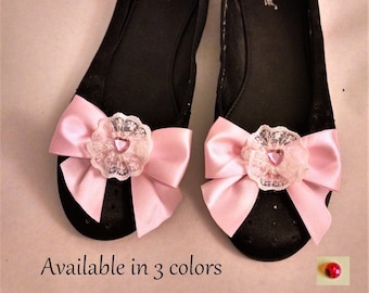 May include: Pair of black ballet flats with pink satin bows and lace accents. Each bow has a small pink heart-shaped rhinestone in the center. The shoes are available in three colors.