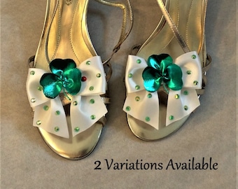 May include: A pair of gold sandals with white bows and green shamrock embellishments. The sandals have a small heel and are decorated with rhinestones. The brand name "LANDOLINO" is visible on the straps.
