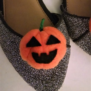 Pode incluir: Um par de sapatos de glitter prata com uma aplica&ccedil;&atilde;o de ab&oacute;bora laranja peluda. A ab&oacute;bora tem um caule, olhos, nariz e boca pretos.