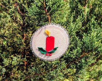 May include: A circular fabric ornament featuring a red candle applique with a yellow flame, accented by green holly leaves and two small red berries. The ornament is edged with white lace trim and hangs from a thin gold cord.