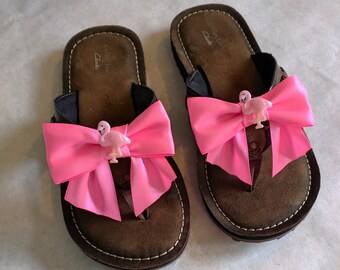 May include: A pair of brown leather sandals with pink bows and flamingo charms. The sandals have the text "Collection Clarks" on the straps.