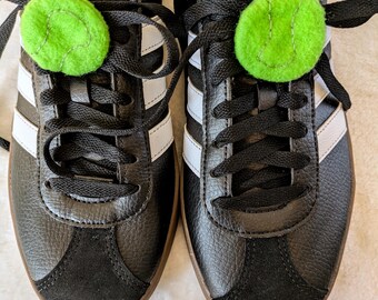 May include: Pair of black Adidas sneakers with white stripes and a green felt ball on the laces. The shoes have black laces and a black suede toe cap. The Adidas logo is visible on the tongue.