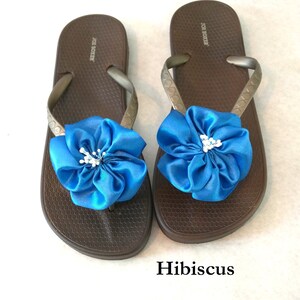 以下が含まれることがあります： メタリックなストラップと、各サンダルに大きな鮮やかな青い布製の花飾りがあしらわれた、茶色のJoe Boxer製ビーチサンダル。フットベッドにはハニカム状のテクスチャーが施されており、サンダルの下にはHibiscusという文字が印刷されています。