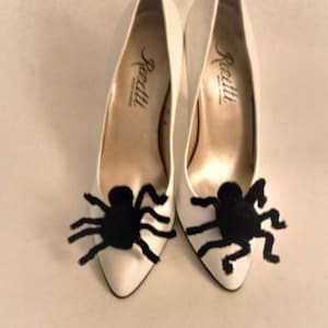 Pode incluir: Um par de sapatos brancos de salto alto com enfeites de aranha peluda preta nos dedos. Os sapatos s&atilde;o etiquetados como "Renatti".